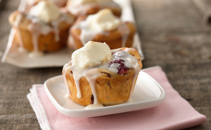 A freshly baked cinnamon roll drizzled with icing and topped with a dollop of cream, served on a small white plate with a pink napkin underneath. More cinnamon rolls are visible on a tray in the background on a wooden surface.